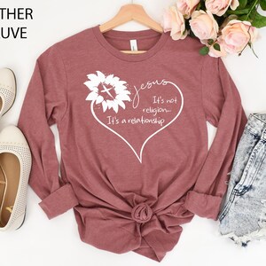May include: A heather mauve long-sleeve t-shirt featuring a white graphic of a sunflower with a cross in the center, shaped into a heart outline with the text: Jesus It's not religion... It's a relationship. The shirt is displayed on a wooden hanger.