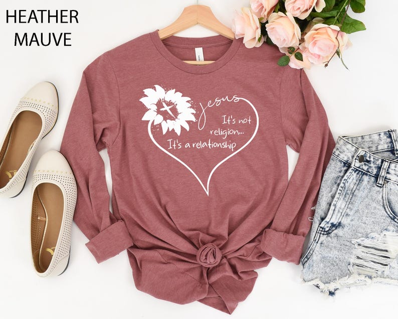 May include: A heather mauve long-sleeve t-shirt featuring a white graphic of a sunflower with a cross in the center, shaped into a heart outline with the text: Jesus It's not religion... It's a relationship. The shirt is displayed on a wooden hanger.