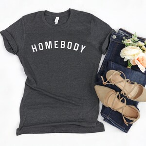Puede incluir: Una camiseta gris oscuro con la palabra "HOMEBODY" impresa en blanco en la parte delantera. La camiseta se combina con unos vaqueros azules, un par de sandalias beige y un ramo de flores blancas y melocot&oacute;n.
