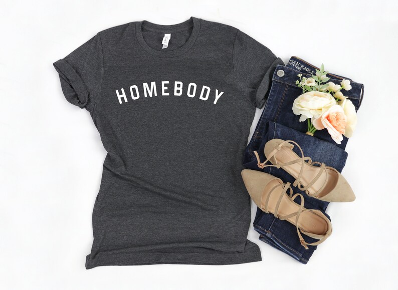 Puede incluir: Una camiseta gris oscuro con la palabra "HOMEBODY" impresa en blanco en la parte delantera. La camiseta se combina con unos vaqueros azules, un par de sandalias beige y un ramo de flores blancas y melocot&oacute;n.