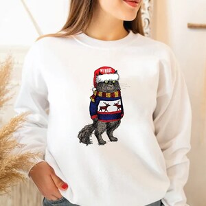 K&ouml;nnte beinhalten: Wei&szlig;er Pullover mit einer schwarzen Katze, die einen blau-wei&szlig;en Pullover mit Rentieren und einer roten Weihnachtsm&uuml;tze tr&auml;gt. Die Katze tr&auml;gt einen gelben und braunen Schal. Der Text "MY MODE" steht auf der Weihnachtsm&uuml;tze.