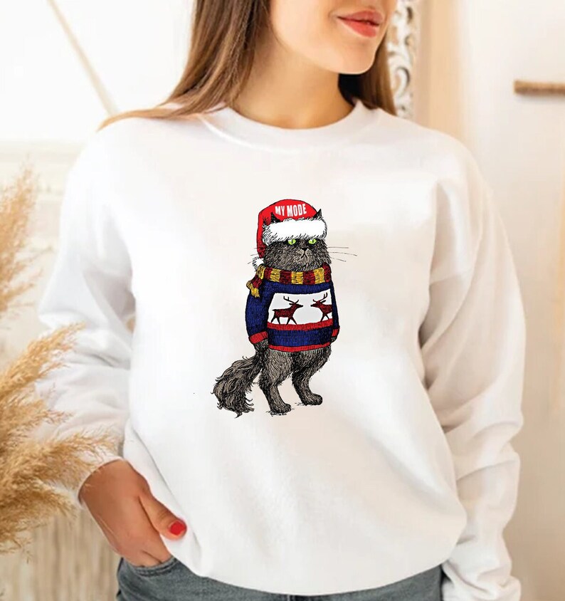 K&ouml;nnte beinhalten: Wei&szlig;er Pullover mit einer schwarzen Katze, die einen blau-wei&szlig;en Pullover mit Rentieren und einer roten Weihnachtsm&uuml;tze tr&auml;gt. Die Katze tr&auml;gt einen gelben und braunen Schal. Der Text "MY MODE" steht auf der Weihnachtsm&uuml;tze.