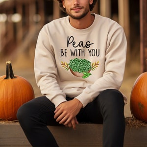 Puede incluir: Una persona lleva una sudadera beige con el texto "Peas Be With You" y un gr&aacute;fico de un taz&oacute;n de guisantes. La persona est&aacute; sentada entre dos calabazas naranjas.