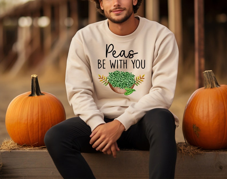 Puede incluir: Una persona lleva una sudadera beige con el texto "Peas Be With You" y un gr&aacute;fico de un taz&oacute;n de guisantes. La persona est&aacute; sentada entre dos calabazas naranjas.