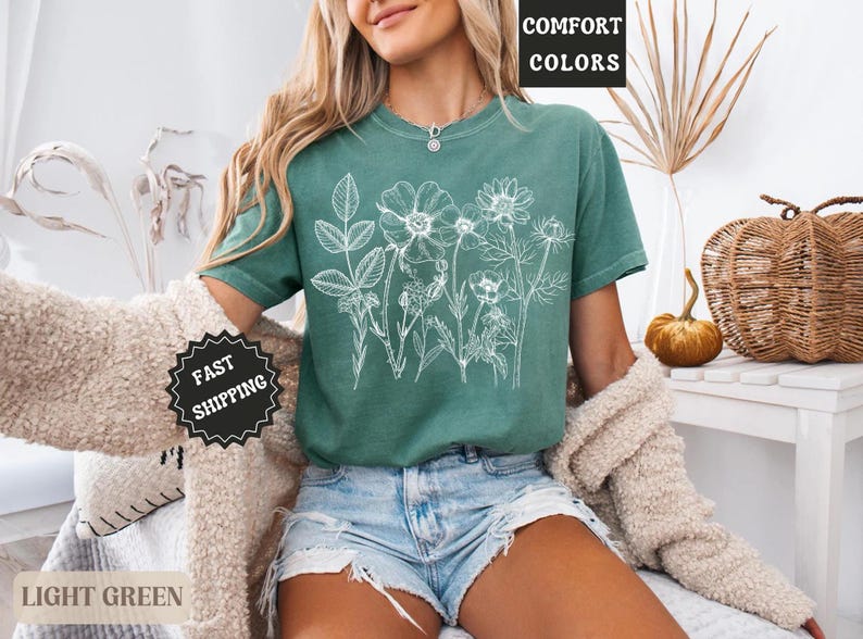 May include: Light green t-shirt with a white floral design. The shirt has short sleeves and a crew neck. The words "COMFORT COLORS" are in a black box in the upper right corner. The words "FAST SHIPPING" are in a black circle.