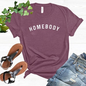 Puede incluir: Una camiseta de color granate con la palabra "HOMEBODY" impresa en blanco en la parte delantera.