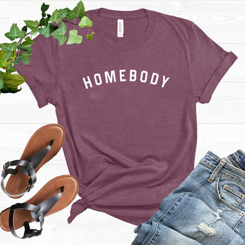 Puede incluir: Una camiseta de color granate con la palabra "HOMEBODY" impresa en blanco en la parte delantera.