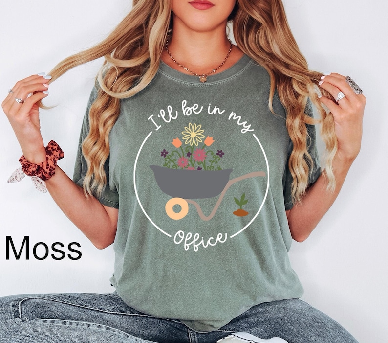 Op de afbeelding: Een groen T-shirt met de tekst "I'll be in my office" in een cirkel rond een kruiwagen gevuld met bloemen.