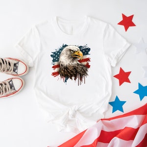 May include: White t-shirt with a graphic of a bald eagle with an American flag in the background. The eagle is looking to the left and has a fierce expression. The flag is red, white, and blue and has a distressed look. The shirt is laying on a white surface with a red, white, and blue American flag in the background.