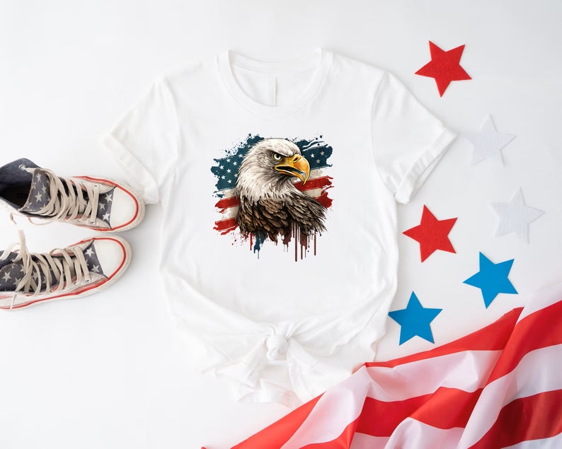 May include: White t-shirt with a graphic of a bald eagle with an American flag in the background. The eagle is looking to the left and has a fierce expression. The flag is red, white, and blue and has a distressed look. The shirt is laying on a white surface with a red, white, and blue American flag in the background.