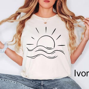 Puede incluir: Camiseta de color marfil con un dibujo lineal negro de un sol saliendo sobre olas oce&aacute;nicas. La camiseta tiene un corte relajado. La palabra "Ivory" est&aacute; impresa en negro en la parte inferior derecha de la camiseta.