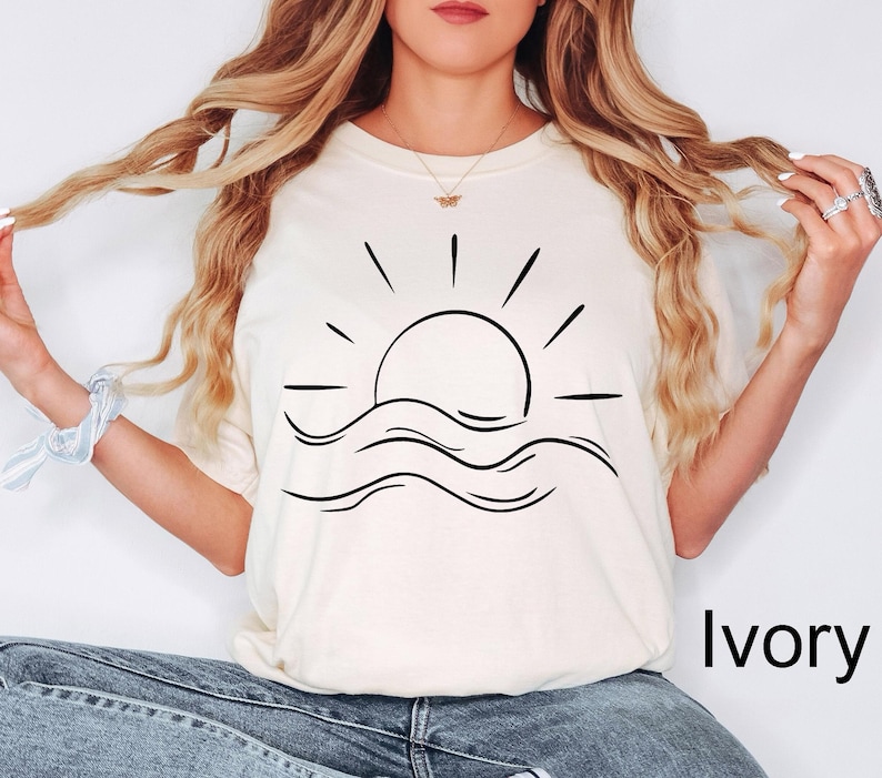 Puede incluir: Camiseta de color marfil con un dibujo lineal negro de un sol saliendo sobre olas oce&aacute;nicas. La camiseta tiene un corte relajado. La palabra "Ivory" est&aacute; impresa en negro en la parte inferior derecha de la camiseta.
