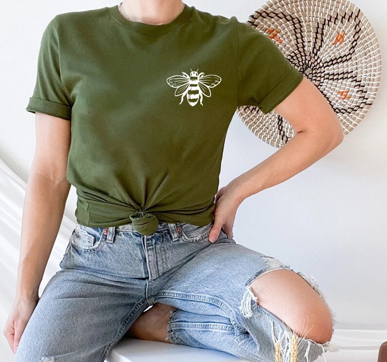 May include: A green t-shirt with a white bee graphic on the front. The shirt is tied at the waist and the model is wearing blue jeans.
