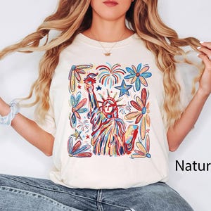 May include: Cream-colored t-shirt featuring a colorful illustration of the Statue of Liberty, fireworks, stars, and flowers. The word "Natural" is printed on the lower right. The design is in red, blue, yellow, and green.