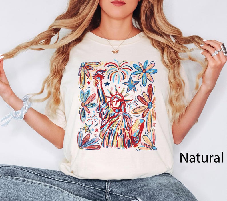 May include: Cream-colored t-shirt featuring a colorful illustration of the Statue of Liberty, fireworks, stars, and flowers. The word "Natural" is printed on the lower right. The design is in red, blue, yellow, and green.