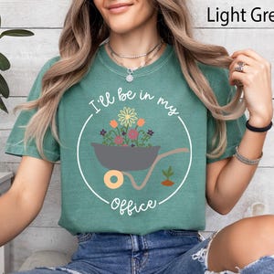 Op de afbeelding: Een lichtgroen T-shirt met een afbeelding van een kruiwagen vol bloemen en de tekst "I'll be in my office".