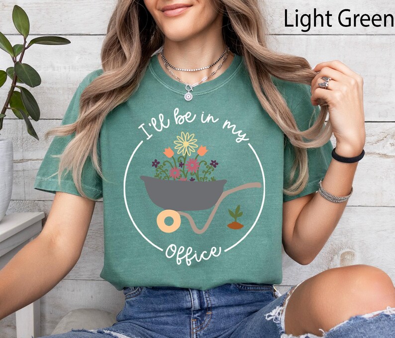 Op de afbeelding: Een lichtgroen T-shirt met een afbeelding van een kruiwagen vol bloemen en de tekst "I'll be in my office".