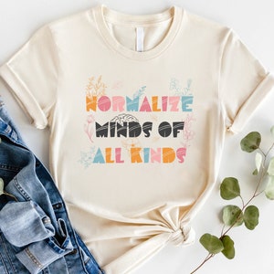 May include: A beige t-shirt with the text "Normalize Minds of All Kinds" in colorful letters with a brain graphic and floral accents.