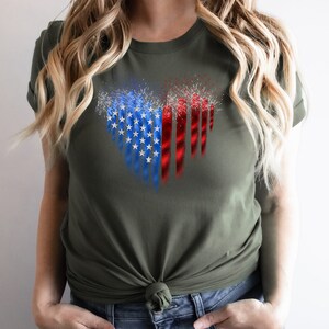 Pu&ograve; includere: T-shirt verde oliva con un design patriottico. La grafica presenta una bandiera americana stilizzata con stelle blu, strisce rosse e fuochi d'artificio bianchi. La maglietta &egrave; realizzata in materiale morbido e ha maniche corte.