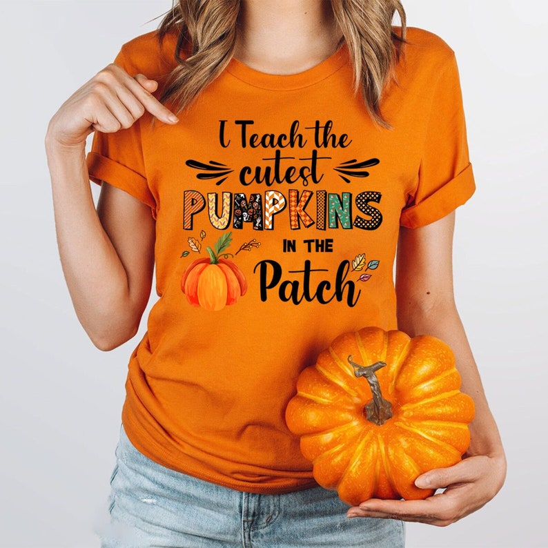 May include: Orange t-shirt with the text "I Teach the Cutest Pumpkins in the Patch." The shirt features a pumpkin graphic and fall leaves. A person is holding a small orange pumpkin.