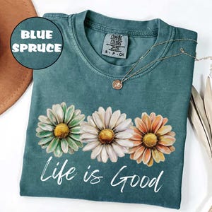 May include: Blue spruce t-shirt with a daisy floral design. The shirt features three daisies in green, white, and orange with yellow centers. The words "Life is Good" are printed in white script below the flowers. A gold necklace is draped on the shirt.