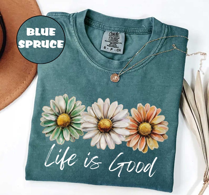 May include: Blue spruce t-shirt with a daisy floral design. The shirt features three daisies in green, white, and orange with yellow centers. The words "Life is Good" are printed in white script below the flowers. A gold necklace is draped on the shirt.