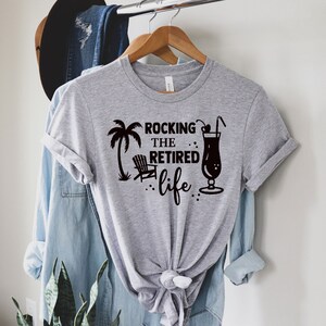 Op de afbeelding: Grijs T-shirt met een zwart grafisch ontwerp met de tekst "Rocking the Retired Life" met een palmboom, een strandstoel en een cocktailglas.