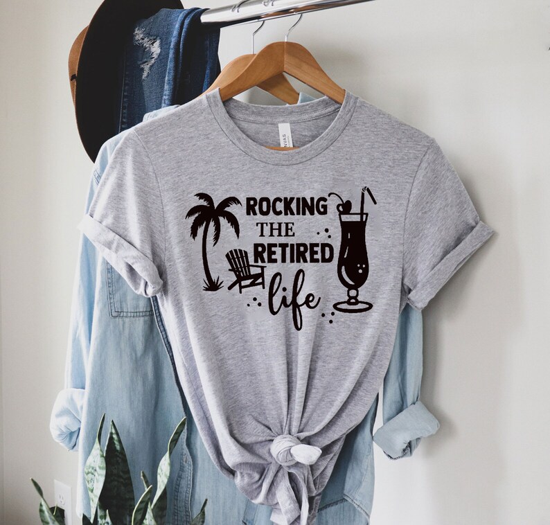 Op de afbeelding: Grijs T-shirt met een zwart grafisch ontwerp met de tekst "Rocking the Retired Life" met een palmboom, een strandstoel en een cocktailglas.