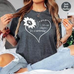 May include: A dark gray heather t-shirt featuring a white graphic of a sunflower and a heart outline. The text reads: Jesus It's not religion... It's a relationship. The design is centered on the chest of the short-sleeved crew neck garment.
