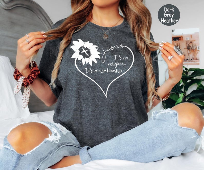May include: A dark gray heather t-shirt featuring a white graphic of a sunflower and a heart outline. The text reads: Jesus It's not religion... It's a relationship. The design is centered on the chest of the short-sleeved crew neck garment.