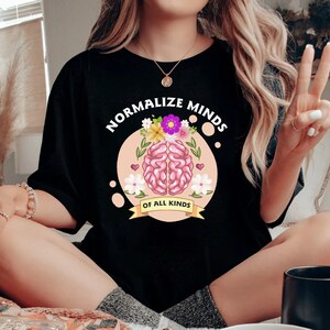 May include: Black t-shirt with a pink brain graphic and the text "Normalize Minds of All Kinds" in white. The brain is surrounded by pink and purple flowers.