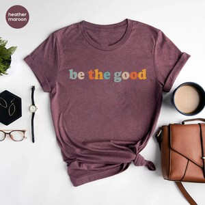 May include: A heather maroon t-shirt with the text "be the good" in colorful letters.