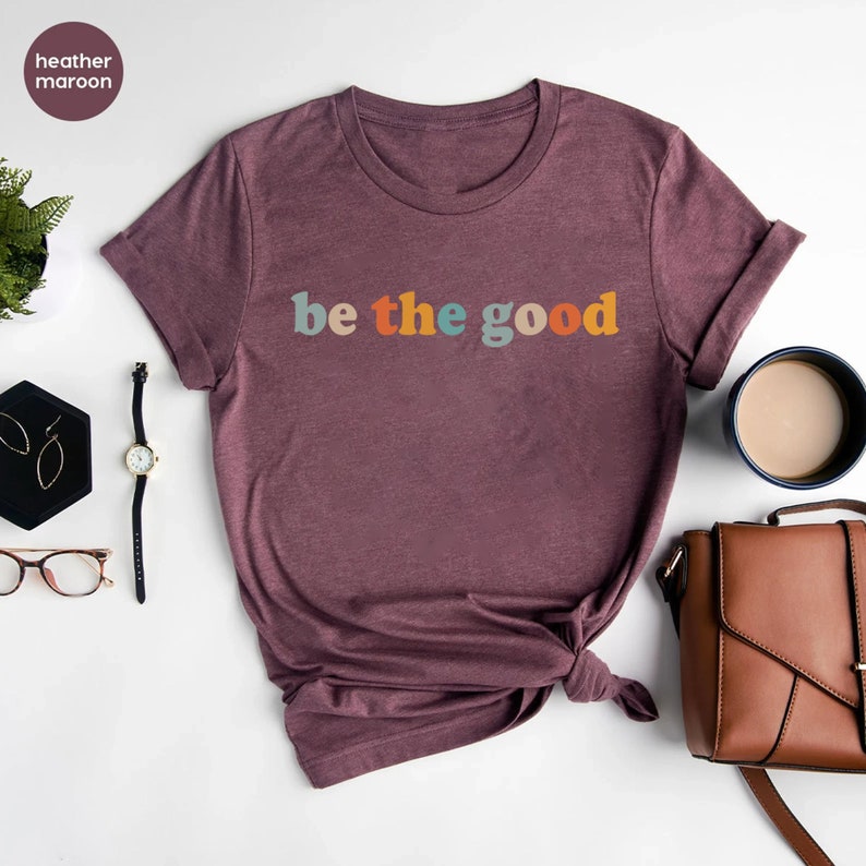 May include: A heather maroon t-shirt with the text "be the good" in colorful letters.