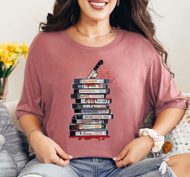 Pode incluir: Camiseta malva com uma pilha de fitas VHS de filmes de terror, uma faca ensanguentada e respingos de sangue. Os t&iacute;tulos incluem "Halloween" e "Hellraiser". Uma pe&ccedil;a casual para os f&atilde;s de filmes de terror.