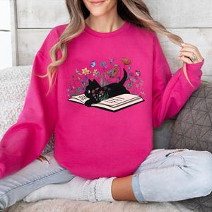 May include: A pink sweatshirt with a black cat reading a book surrounded by flowers. The cat is sitting on an open book with the text "Read" on the page.