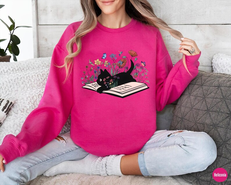 May include: A pink sweatshirt with a black cat reading a book surrounded by flowers. The cat is sitting on an open book with the text "Read" on the page.