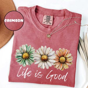 May include: Crimson-colored t-shirt with three daisy flower graphics and the words "Life is Good" in white script. A silver necklace with a pendant rests on the shirt. The shirt is a Comfort Colors brand.