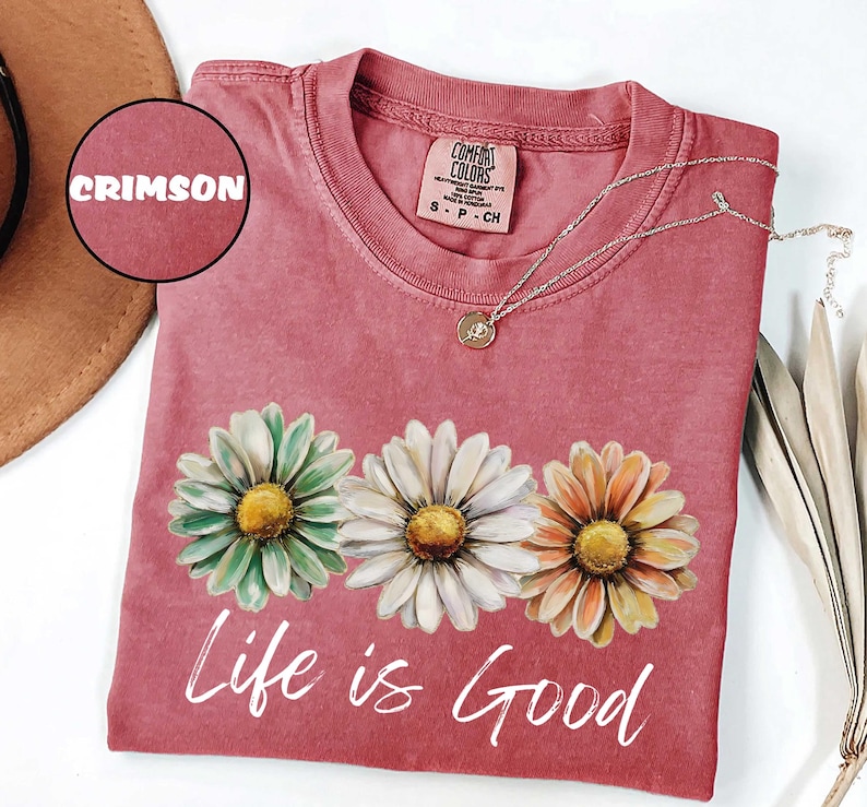 May include: Crimson-colored t-shirt with three daisy flower graphics and the words "Life is Good" in white script. A silver necklace with a pendant rests on the shirt. The shirt is a Comfort Colors brand.