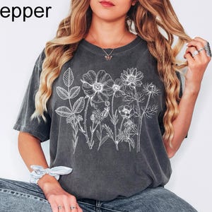 May include: Dark gray t-shirt with a white floral design. The shirt features a botanical illustration of various flowers and leaves. The word "Pepper" is printed in the upper left corner.