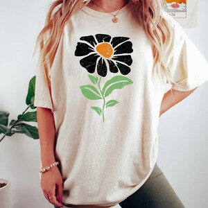 May include: A cream colored t-shirt with a black and orange flower graphic. The flower has a green stem and leaves.