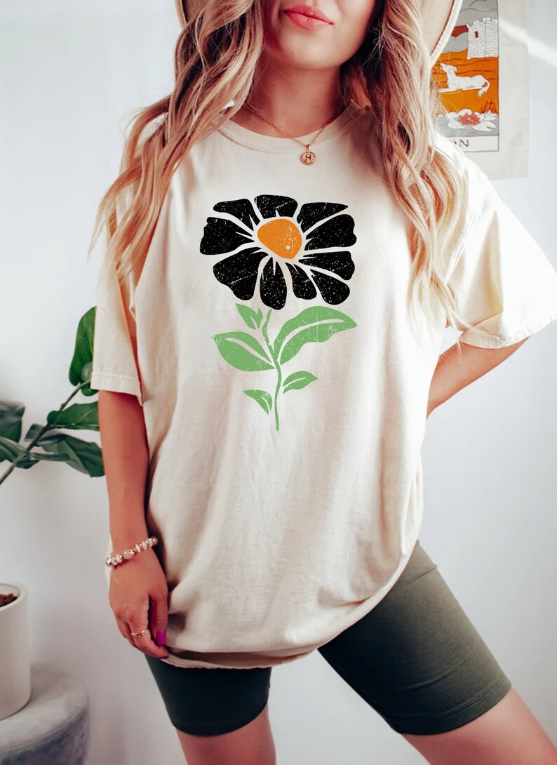 May include: A cream colored t-shirt with a black and orange flower graphic. The flower has a green stem and leaves.