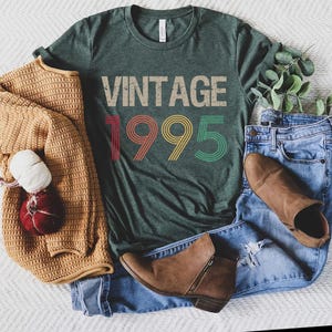 May include: A green t-shirt with the text "Vintage 1995" in rainbow colors. The shirt is laid out on a white surface with a brown sweater, blue jeans, and brown boots.