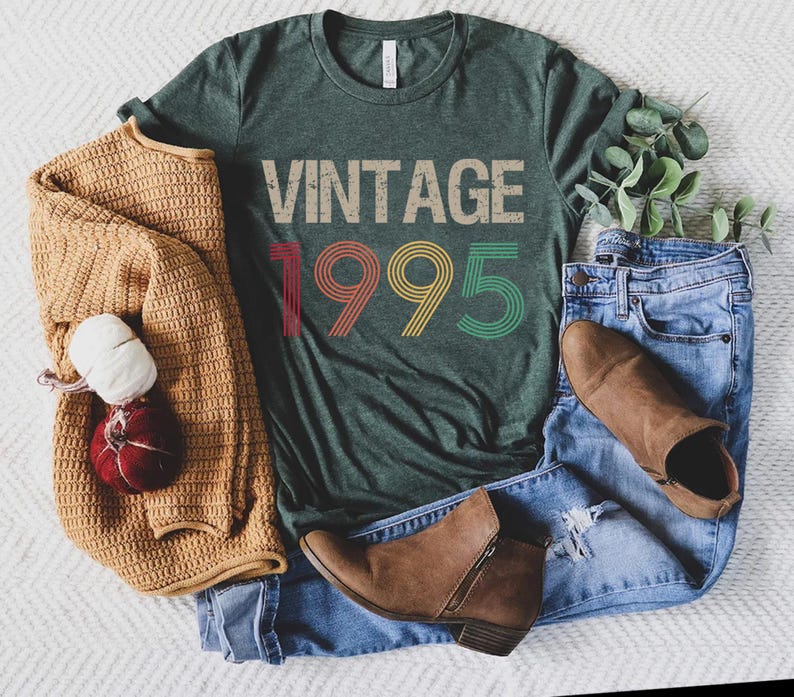 May include: A green t-shirt with the text "Vintage 1995" in rainbow colors. The shirt is laid out on a white surface with a brown sweater, blue jeans, and brown boots.