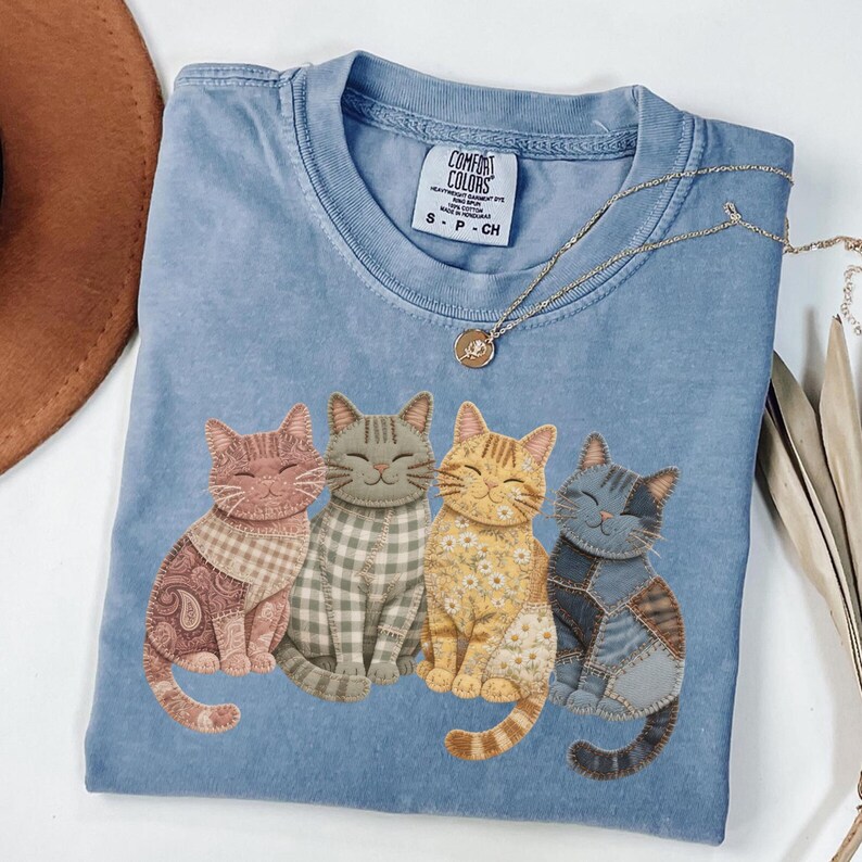 May include: A light blue Comfort Colors t-shirt featuring four smiling cat illustrations. Each cat has a unique patchwork design in shades of pink, gray, yellow, and blue. A gold necklace with a pendant rests on the shirt.