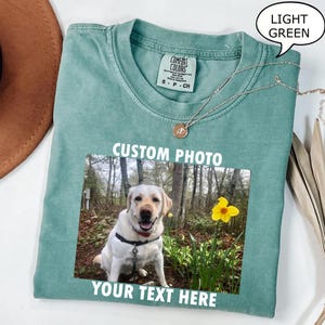 Peut inclure: T-shirt vert clair avec une photo d'un labrador jaune et d'une jonquille. Le t-shirt porte les mots "CUSTOM PHOTO" et "YOUR TEXT HERE". Un collier argenté avec un pendentif est drapé sur le t-shirt.