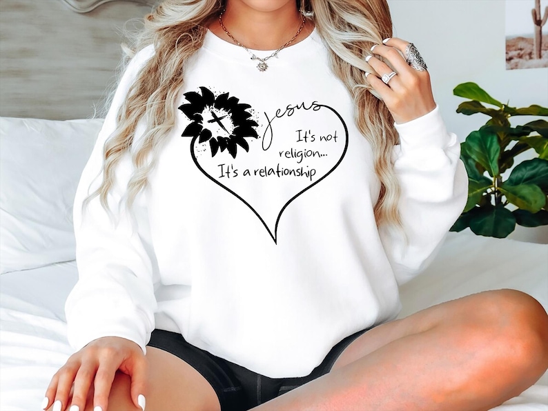 May include: A white long-sleeve sweatshirt featuring a black graphic print of a sunflower with a cross in the center, shaped into a heart outline. The text reads: Jesus It's not religion... It's a relationship, written in a cursive and print font.
