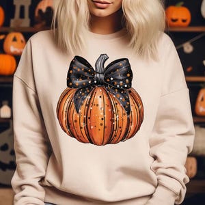 May include: A beige sweatshirt featuring a graphic of an orange pumpkin with a black bow and gold glitter. The pumpkin has a black stem and black stripes.