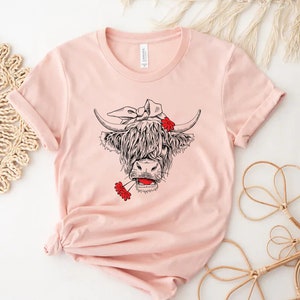 May include: A pink t-shirt with a black and white illustration of a Highland cow wearing a red bandana and holding red flowers in its mouth.