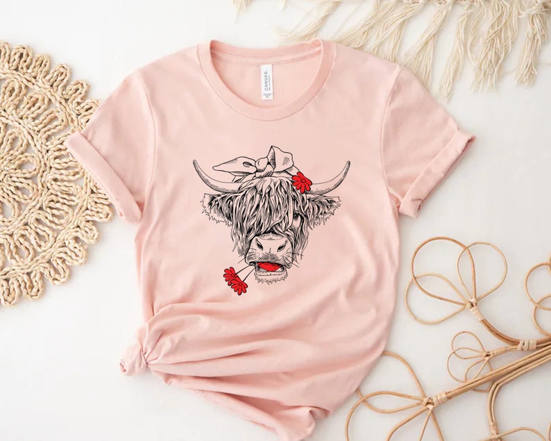 May include: A pink t-shirt with a black and white illustration of a Highland cow wearing a red bandana and holding red flowers in its mouth.