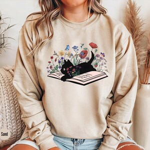 May include: A beige sweatshirt with a black cat reading a book surrounded by flowers. The cat is lying on the open book. The flowers are in shades of pink, purple, blue, and yellow.